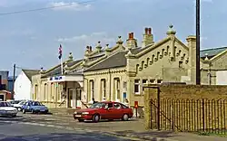 A photograph of a brick building against a blue sky taken from a short distance away. Two cars are parked in front and a sign on top of a canopy coming out from the building says King's Lynn to the right of a double arrow logo. A Union Flag is flying atop the canopy.