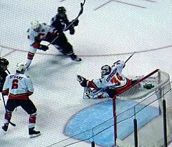 View from behind the left shoulder of a goaltender as he reaches out with his left hand and catches a puck shot into his glove as several players look on.