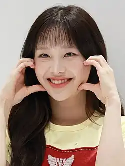 A young Asian woman with long brown hair and a fringe wearing a yellow t-shirt grinning widely with her fingers and thumbs on each of her cheeks as if forming a heart.