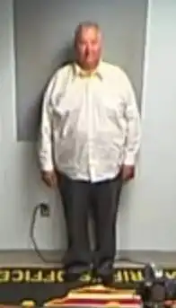 A full-body length image of Householder in a white suit against a gray wall