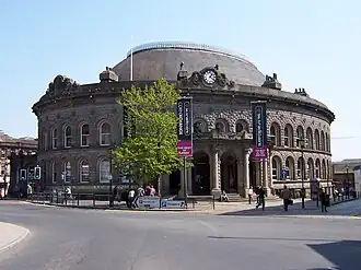 Photograph of a large stone building with an oval footprint and a domed roof. The main entrance lies behind four large arches and underneath a clock. Numerous arched windows lie on two levels all around the building.