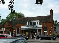 A small red-brick station building as viewed from across the street. There is a entranceway with a canopy and dormer window above; on this canopy is a sign reading "Letchworth Garden City". On either side of the central section the canopy merges into a bay window. The station is fairly crowded in front with people and cars.