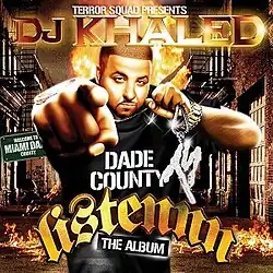 A man wearing a black t-shirt saying "Dade County" while holding a platinum "Terror Squad" necklace, points at the viewer while a fire happens behind him.
