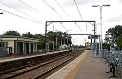 -A view of a two-track electrified railway from the right-hand platform. At the end of the platform there is a closed level crossing gate and a cream and green signal box. On the left-hand platform there is a small creeam and green shelter.