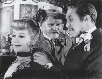 young, white, moustached man in Edwardian clothes putting a necklace on a young white woman, watched by a whiskered older man in a straw hat