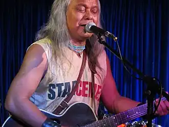 Lou Majaw wearing a light-coloured, sleeveless print t-shirt, playing acoustic guitar and singing into a microphone onstage