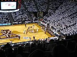 a crowd of people in mostly white T-shirts watch an indoor basketball game; a T with a start on it is upside down and featured prominently under the players