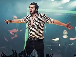 A dark haired man wearing a striped shirt extends his arms to the crowd while performing with a microphone