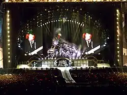 AC/DC shown on stage with huge screens either side depicting Angus on guitar. He is actually at extreme stage right the rest of the band at centre stage. They are beneath a huge steam engine, tilted slightly upwards, with AC/DC's logo on its side.
