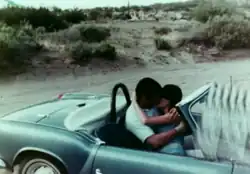 A man and woman kiss in a convertible sports car, with the clapperboard accidentally at the edge of the frame.