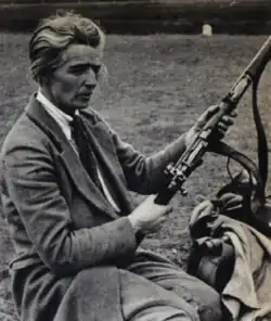 Foster, seated, in a tweed jacket, blouse and tie, holding a bolt-action target rifle with a sling fitted