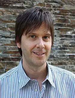 alt=Portrait photo of a white man with dark brown hair wearing a striped dress shirt.