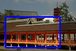 A roofed corridor and two connected buildings with gabled roofs. All of them have vermillion red wooden beams, white walls and are standing on stilts. Beyond the buildings in the background on a hill stands a five-storied red colored pagoda and a large building with gabled roof.
