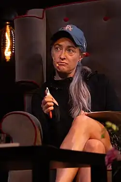 Matylda Damięcka sitting in a chair with legs crossed, wearing a black hoodie, dark blue print baseball cap, and a headset microphone, pen in one hand, looking right of camera with pursed lips