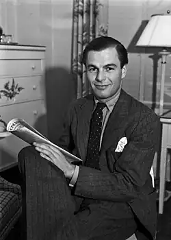 A Caucasian man wearing a suit, sitting on a chair with legs crossed, facing slightly left, and holding a newspaper in his lap