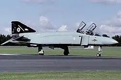 Side view of a Phantom F.3 with both cockpit canopies open. the aircraft is in air superiority grey with a black tail, and has a tiger's head painted on the nose, and a tiger's head in a white disc on the tail. These markings represent Seventy-four Squadron