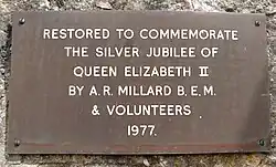 Colour photograph of the brass plaque with the commemoration written in upper case on white lettering. The commemoration reads as follows: "Restored to commemorate the Silver Jubilee of Queen Elizabeth II by A. R. Millard B.E.M. & volunteers 1977."