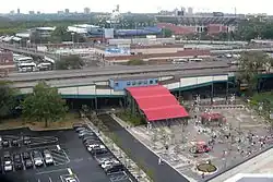The entrance/exit stairs for the Mets-Willets Point station as viewed from Citi Field. The stairs are extremely wide and are covered by a red canopy, which leads from a plaza in the foreground to the station's mezzanine in the background. The Long Island Rail Road station is at rear left and the USTA National Tennis Center is at rear right.