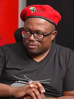 A man seated at a table, wearing glasses, a red beret and black v-neck t-shirt