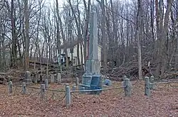 A group of graves inside a small metal railing low to the ground. In the middle is a large gray obelisk. Behind are bare trees, through which a nearby yellow house is visible.