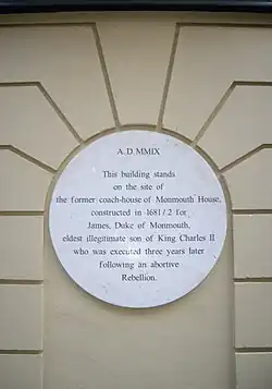 A white plaque on a beige wall noting where Monmouth House stood in Soho Square