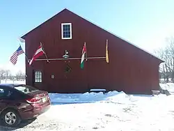 A red barn displaying the flags of Austria, Hungary, New Jersey, and the United States, with snow on the ground and a car in front.