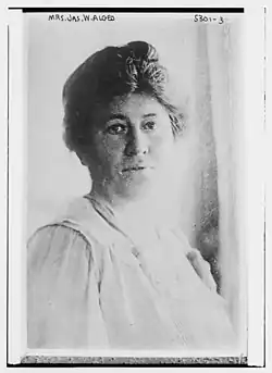 A white woman with dark hair in a simple updo. She is wearing a light colored dress.