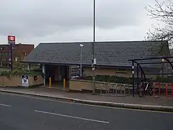A small yellow-brick station building that is mostly covered by its slate-tile roof as viewed from across the street. The centre of the building is cut out to be a gateway but the door arch is closed by a gate. To the left there is a blue pole with a double arrow logo on top and a sign that reads "New Barnet".