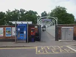 The entrance to a cast iron footbridge with a tall brick wall on either side, as photographed from across the road. There are cast iron spans overhead between the spans, and on the front one there is a sign that reads "New Southgate". On the left of the entrance there is a ticket machine and other posters.