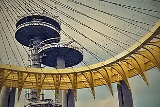 The outer and inner girders of the Tent of Tomorrow, which are curved and painted yellow. There are wires extending inward from the girders. The observation towers are in the background.