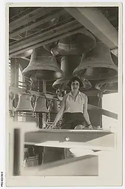 A woman with short, brown hair sitting on a wooden beam with large bells hanging in the background