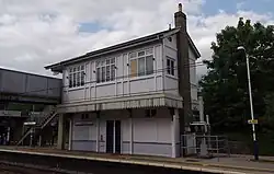 a two-storey white-painted wood building with brick highlights. The building is designed such that the bottom storey is on a railway island platform, and the upper storey is on the footbridge above. Part of the footbridge, stairs up to it on the opposite side, overhead wires, and a lamppost are also visible.