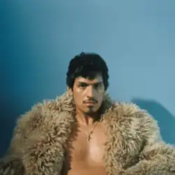 Omar sitting partially bare-chested, wearing a furry jacket and a cross necklace, looking ahead at the camera in front of a blue background.