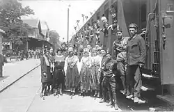 A group of men and women posing for a photo, some wearing uniforms or folk costumes, standing on a railway platform beside a train carriage fron which more people are leaning outside the carriage windows and doors looking at the camera