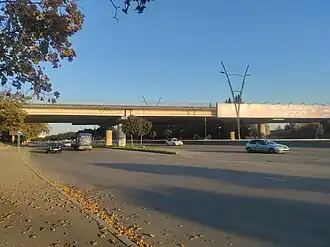 A wide shot of a concrete overpass spanning a multi-lane road on a sunny day. Cars are visible on the road below, and autumn leaves litter the sidewalk in the foreground.