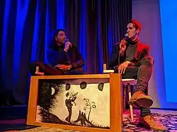 Two young men sitting on chairs on a stage holding microphones, with a crankie in front of them, depicting a fiddler