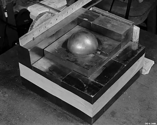 A stack of square metal plates with a side about 10 inches. In the 3-inch hole in the top plate there is a gray metal ball simulating Pu.