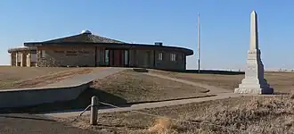 Low circular stone building with low-pitched conical roof; square granite obelisk in foreground