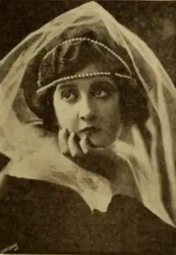 A young white woman with dark hair and eyes, wearing a headpiece with pearl bands and tulle, one hand on her cheek