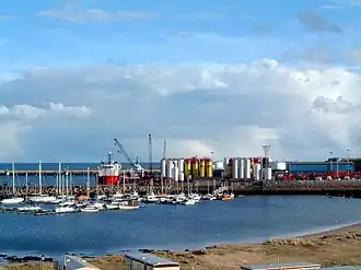 Peterhead Bay. In the background the breakwater built by convict labour. In the middle distance silos of drilling mud for the offshore oil industry and yachts berthed in the Peterhead marina. In the foreground the roofs of holiday caravans and the "Lido" sands.