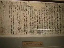 A tattered, ripped and crumpled piece of light coloured paper with writings penned in Chinese chearacters in dark ink, pinned on a wall for display in a museum.