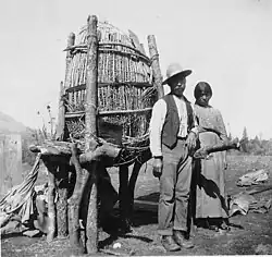 A man and woman of the Mono tribe stand in front of an acorn cache, similar to a large woven basket held up by thick wooden sticks
