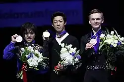 A photograph of Shoma Uno, Nathan Chen and Mikhail Kolyada (from left to right) posing with medals around their necks and flowers in their hands.
