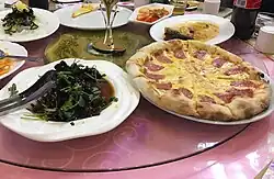 Seven dishes, including a pizza with a slice of meat on each slice, served on a round table.
