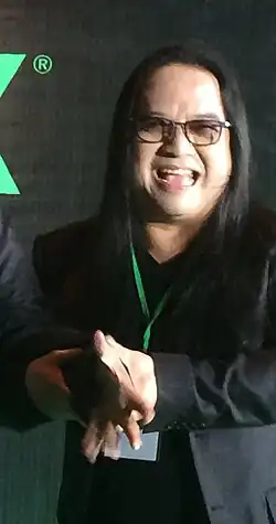 cropped image of Pathompong Sombatpiboon wearing a dark suit and a green lanyard, laughing at camera and appearing to be pulling the hand of someone off-camera