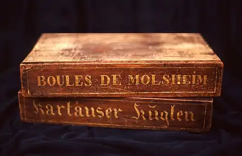 Two wooden boxes on top of each other, on which are written "Boules de Molsheim" and "Kartäuser Küglen" respectively.