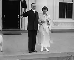 Calvin stands on the pavement in a suit waving his hat. Grace stands next to him smiling in a coat.