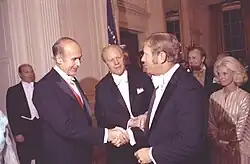 "Two men in formal wear, one the President of France and one President of the United States, greet a third man in formal wear during a state dinner party."