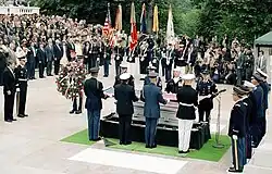 Four men, in military uniforms with different combinations of white and blue, stand on either side of a coffin at lower right, out of doors on a stone floor. Behind them are a group of flags. In the background and at left is an audience on risers