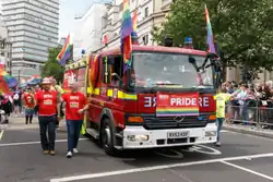 Two firefighters in red "LFB PRIDE" t-shirts walk in a parade alongside a fire engine. The fire engine is decorated with at least five rainbow flags.
